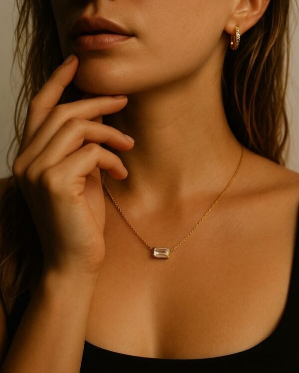 Personne avec un collier en or, pendentif rectangulaire transparent, boucles d'oreilles dorées.